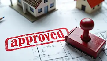 Red "Approved" rubber stamp on architectural blueprint with house model in background.