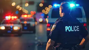 Police officer standing on wet street at night with emergency vehicle lights flashing in background.