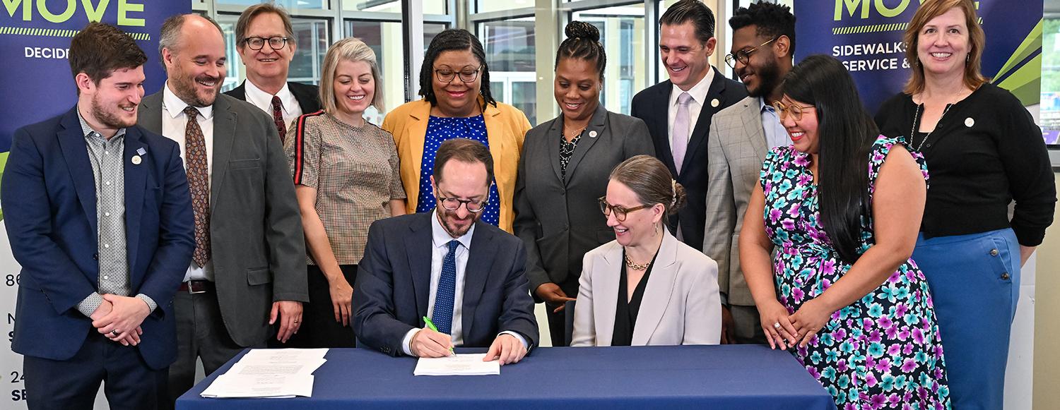 photo of a group of people watching Mayor O'Connell sign a document.