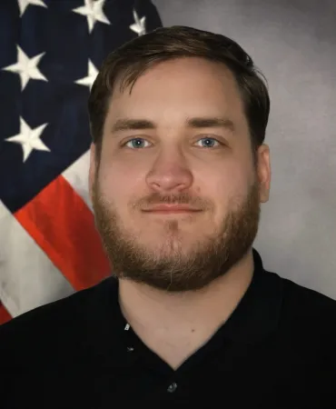 Man with beard posing in front of American flag backdrop for official portrait.