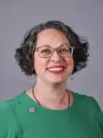 Woman wearing glasses and green shirt smiling at camera against gray background.