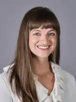 Woman with long brown hair and bangs smiling at camera, wearing white collared shirt against gray background.